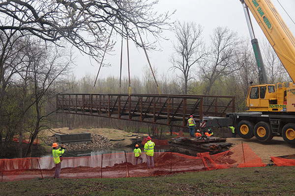 Crane holding bridge span over water with construction workers watching